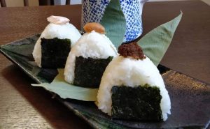 Tre onigiri giapponesi in occasione di festival giapponesi 2019 a roma e milano|Preparazione del sushi con il tappetino in bambù per compattare il riso||Una donna in abito giapponese serve sake in bottiglia|Sushi