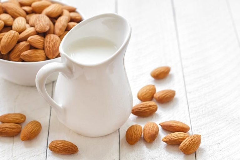 latte di mandorla in bricco bianco con mandorle e ciotola di mandorle su tavolo a doghe in legno bianco