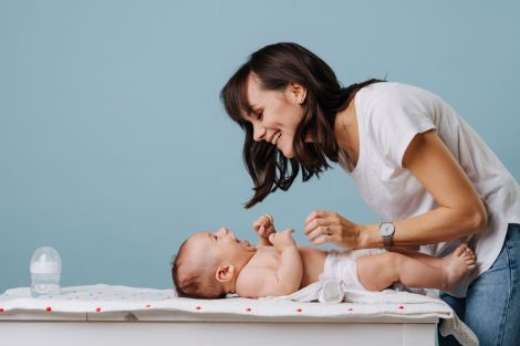 Mother changing diaper on her baby on table over blue background. They look at each other, smile and laugh.