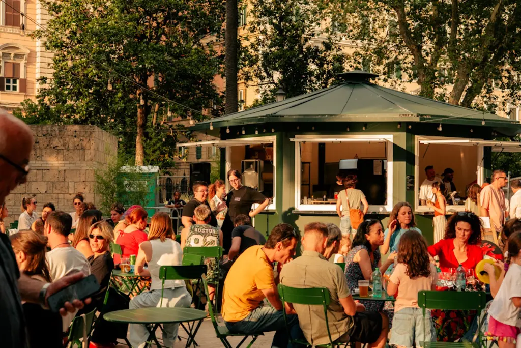 Persone sedute ai tavolini sotto gli alberi di Piazza Vittorio durante una serata estiva a Roma.