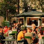 Persone sedute ai tavolini sotto gli alberi di Piazza Vittorio durante una serata estiva a Roma.