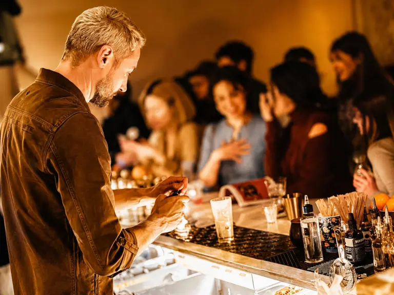 Bancone di cocktail bar affollato ai Navigli di Milano con bartender al lavoro e drink colorati serviti ai clienti