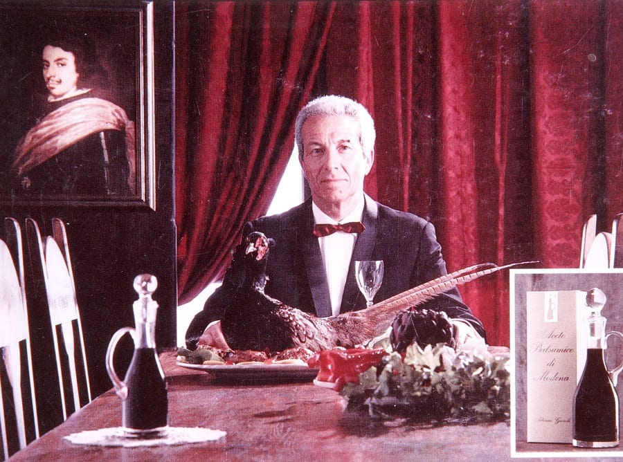 Pubblicità di Adriano Grosoli per l'aceto balsamico, a tavola vestito in frac e papillon, con l'aceto basamico e una tenza di velluto rosso alle spalle