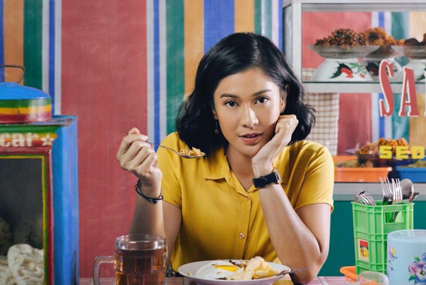 Una donna indonesiana con la camicetta gialla mangia un piatto di pollo. Sullo sfondo una parete colorata, con righe verticali rosse, blu, verdi e arancioni