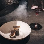 Un dessert di coda, con cloche nera che scopre l'affumicature e bicchiere di vino, su tavolo scuro