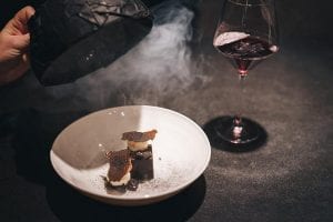 Un dessert di coda, con cloche nera che scopre l'affumicature e bicchiere di vino, su tavolo scuro