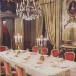 La sala di Palazzo Saluzzo con la tavola apparecchiata, con candelabri, stoviglie di porcellana, sedie in velluto rosso, un lampadario a gocce e tendaggi oro sullo sfondo
